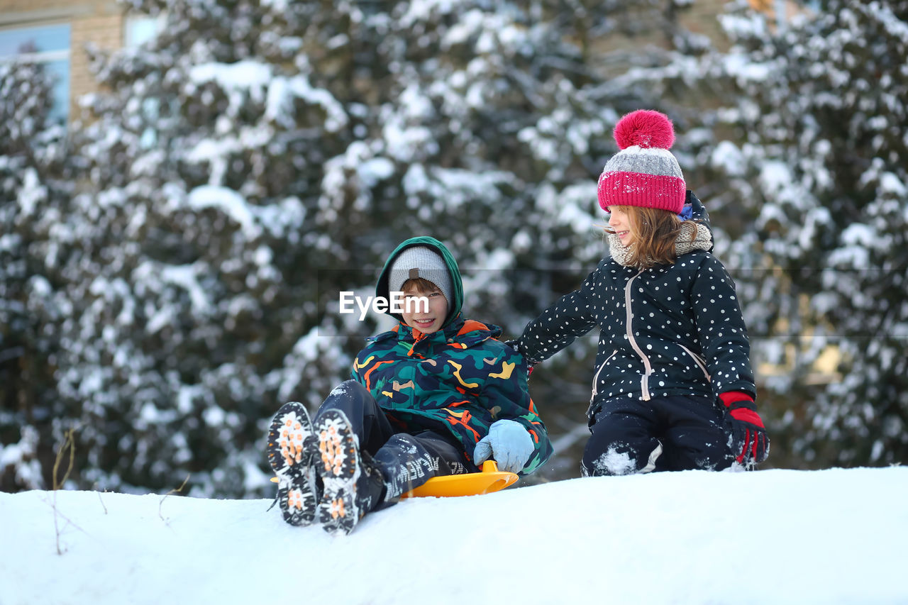 Winter portrait of children with a plastic sled sliding on a snowy slope and making a snowman