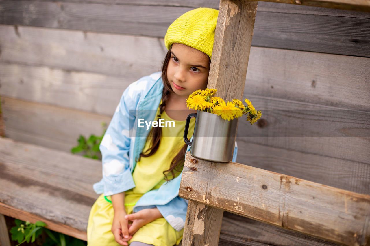 Portrait of a sad adorable little girl in a yellow beret, blue raincoat with bouquet dandelions