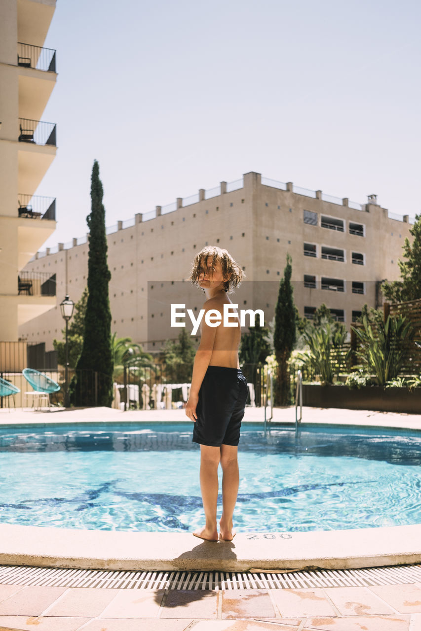 Full length of shirtless boy standing by swimming pool against sky in resort