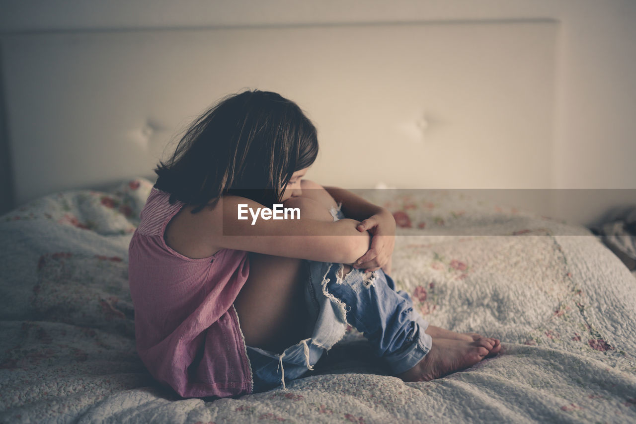 Girl child serious and frustrated sitting on a bed in a bright bedroom, concept age crisis 