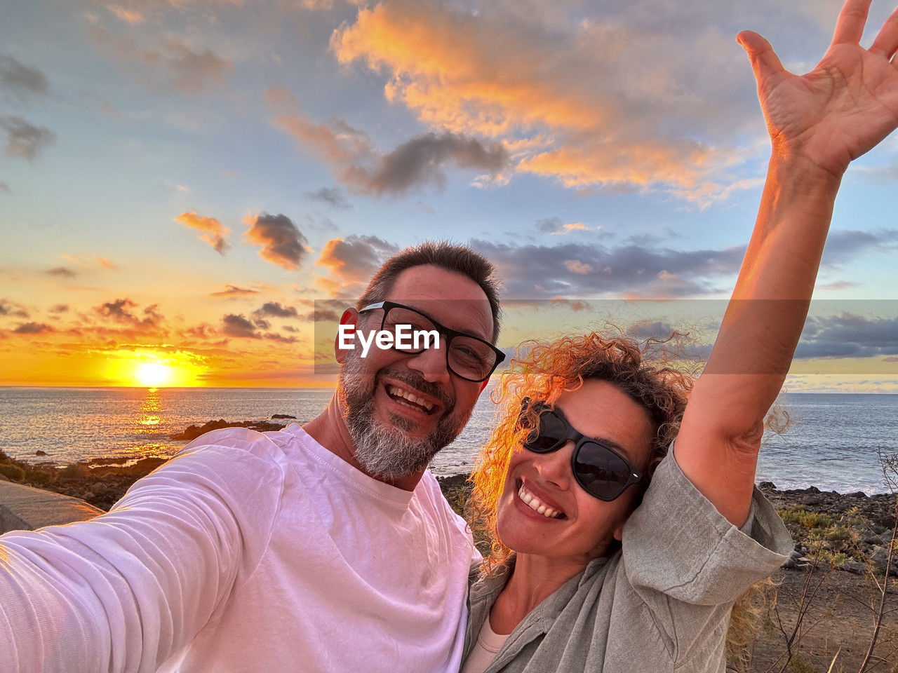 portrait of happy friends with arms raised standing at beach against sky during sunset