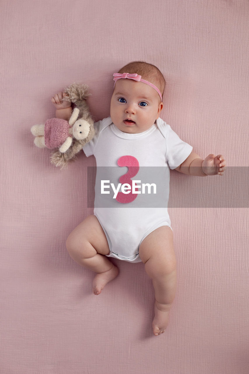 Girl infant with a bow lies on the bed in a white bodysuit with a doll toy child has a pink number 3