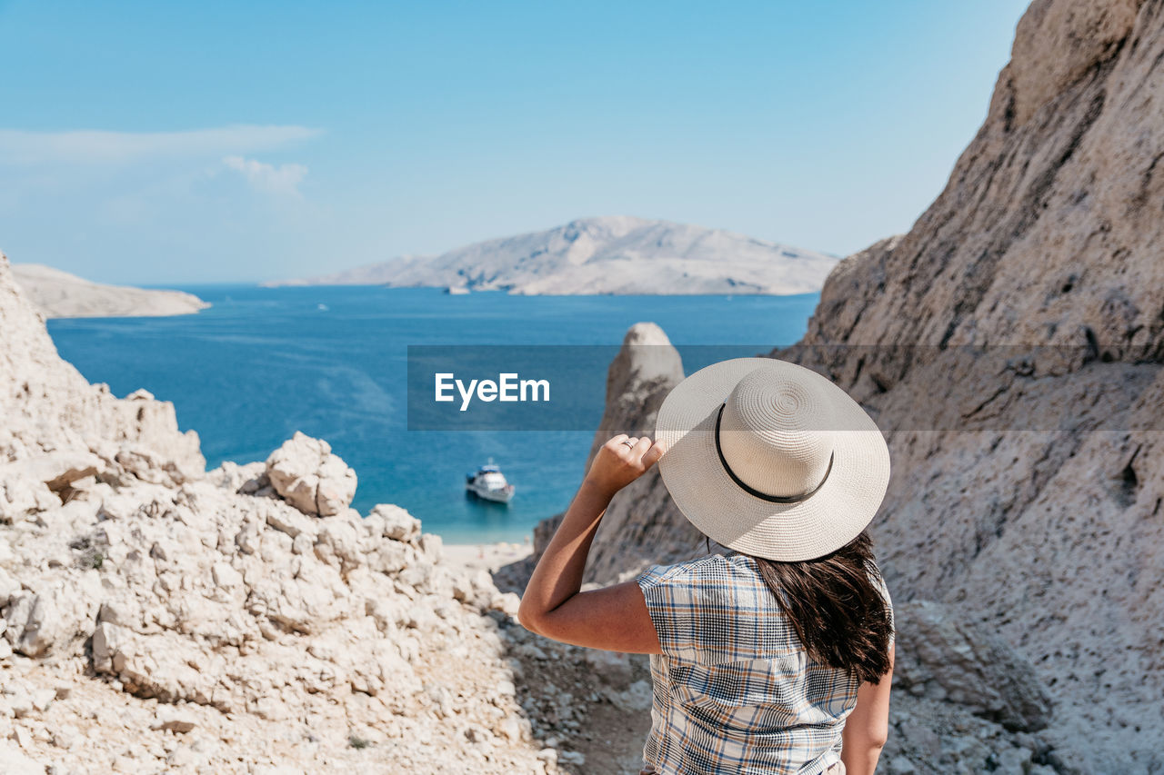Rear view of woman in summer clothes and sun hat looking at spectacular view and sea coastline.