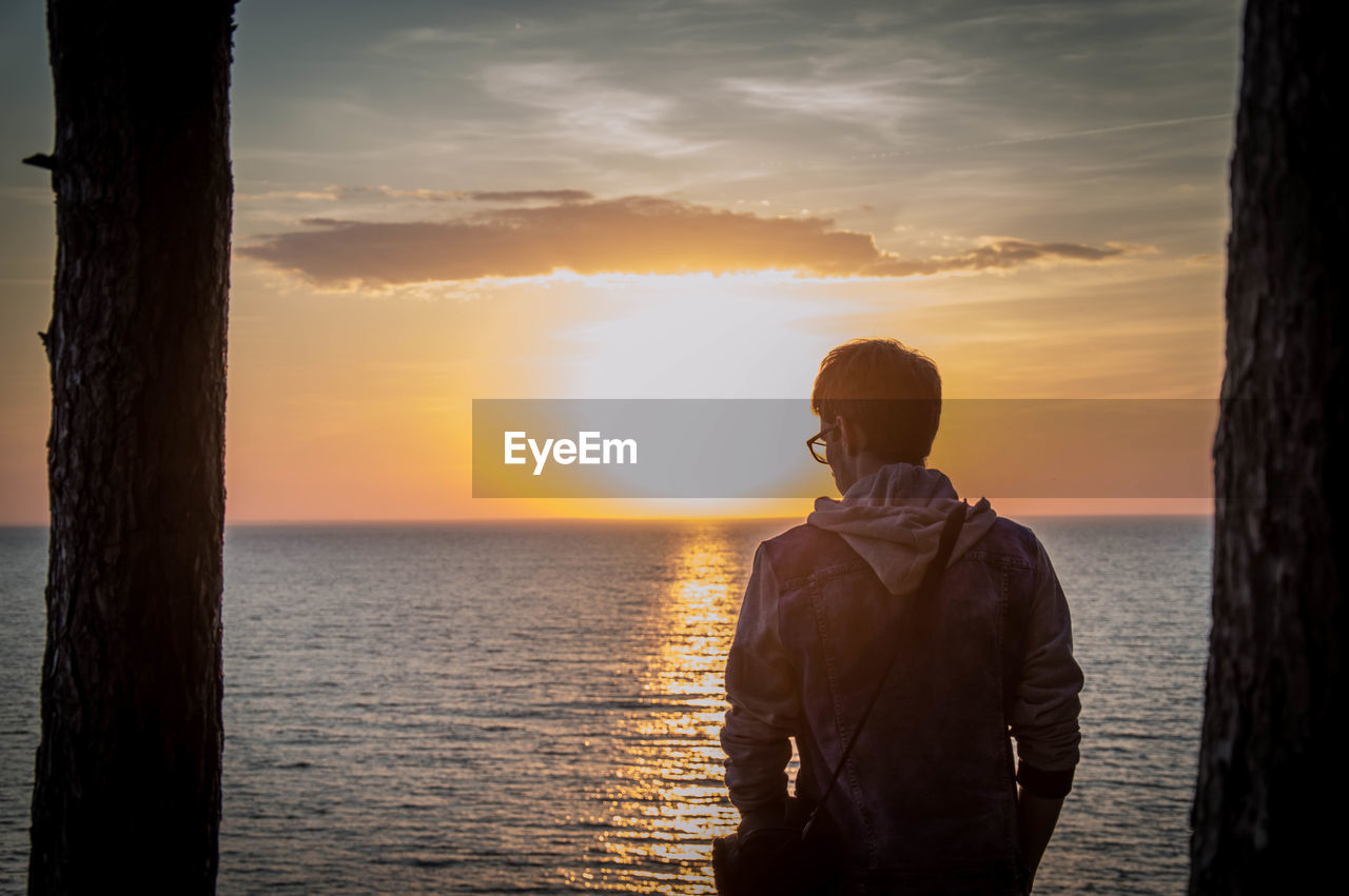 Rear view of man standing by sea against sky during sunset