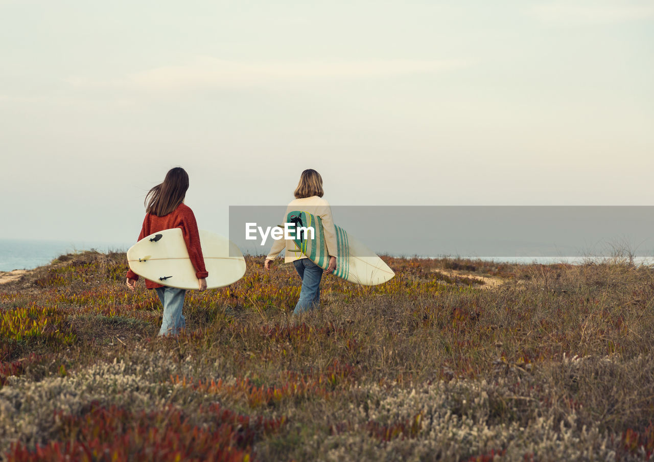 Rear view of women with surfboard walking by plants against sky