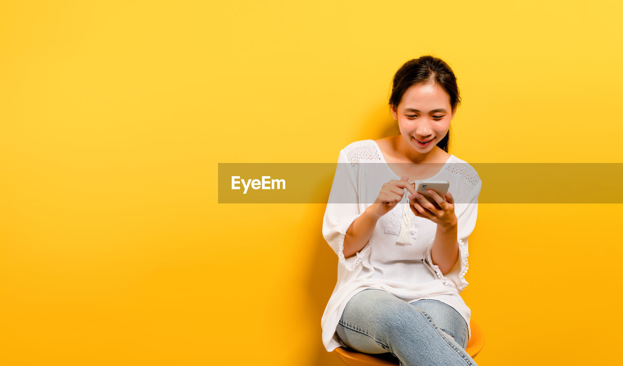 YOUNG WOMAN USING MOBILE PHONE AGAINST YELLOW WALL