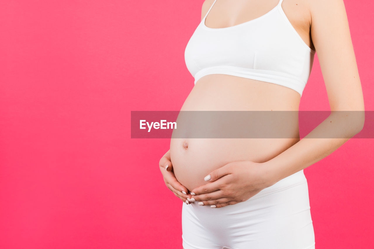 MIDSECTION OF YOUNG WOMAN STANDING AGAINST PINK BACKGROUND