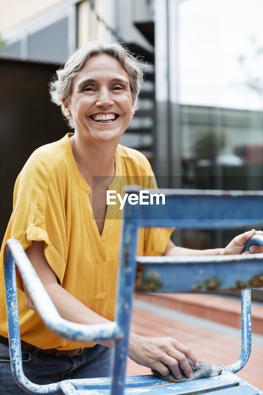 Portrait of cheerful mature woman scrubbing metallic chair with steel wool at yard