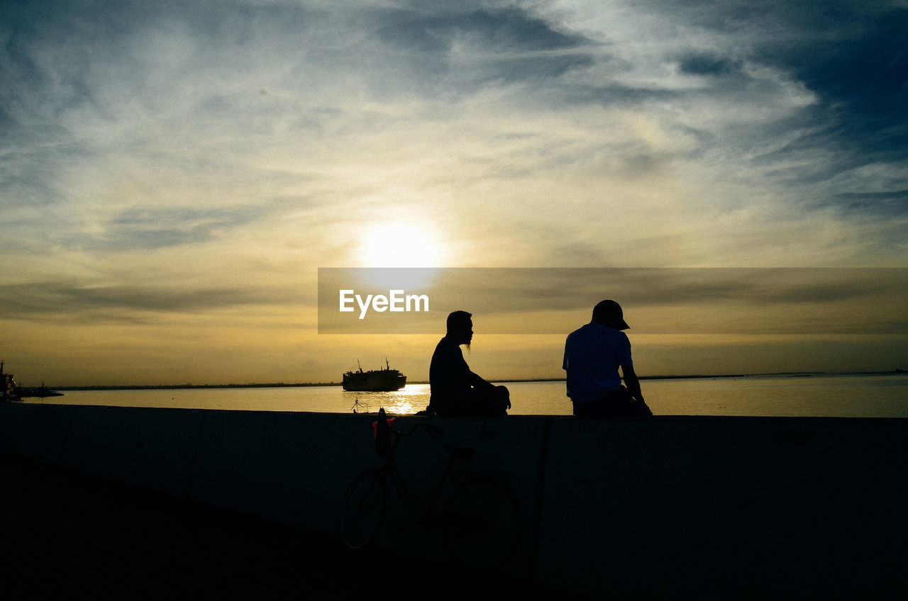 SILHOUETTE MEN SITTING ON SEA AGAINST SKY DURING SUNSET