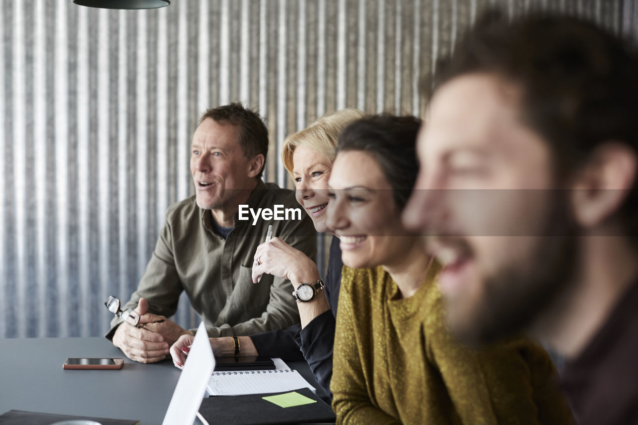 Smiling creative business colleagues sitting at conference table while looking away in board room