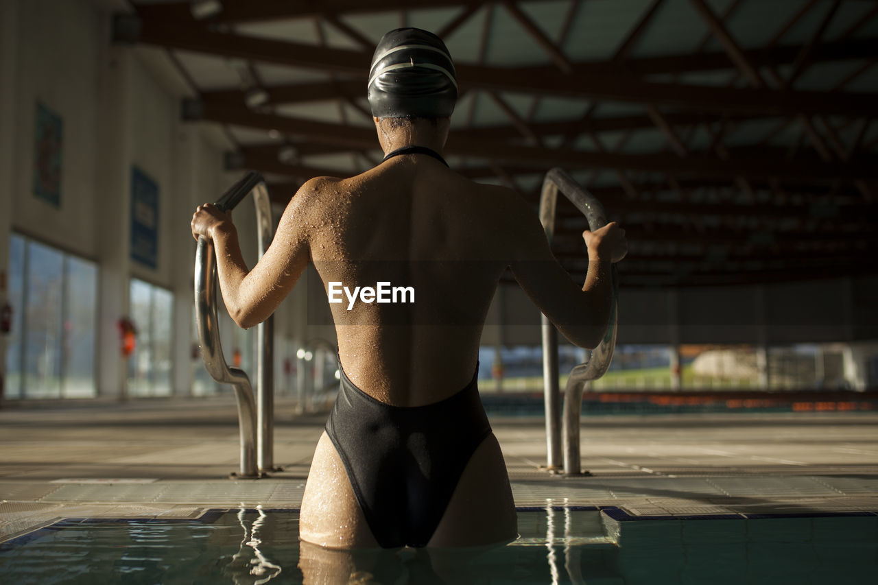 Young beautiful woman leaves the indoor pool by the stairs with black swimsuit, rear view