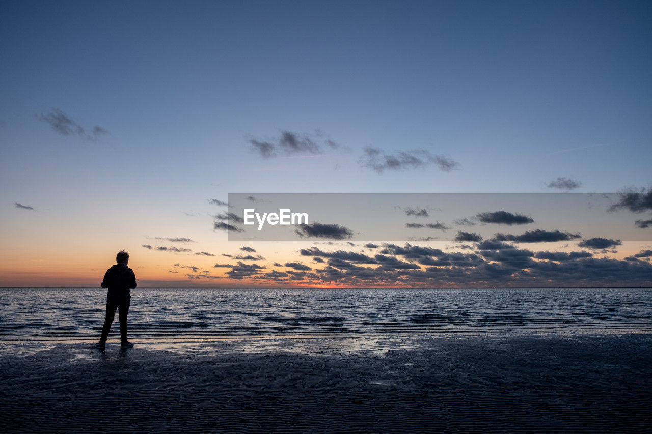 sky, horizon, sea, ocean, water, sunset, cloud, land, beach, one person, nature, wave, beauty in nature, dawn, full length, scenics - nature, shore, silhouette, coast, body of water, adult, holiday, standing, evening, vacation, trip, travel destinations, landscape, tranquility, travel, men, leisure activity, environment, sunlight, horizon over water, reflection, outdoors, tranquil scene, solitude, lifestyles, motion, sports, blue, copy space, walking, activity
