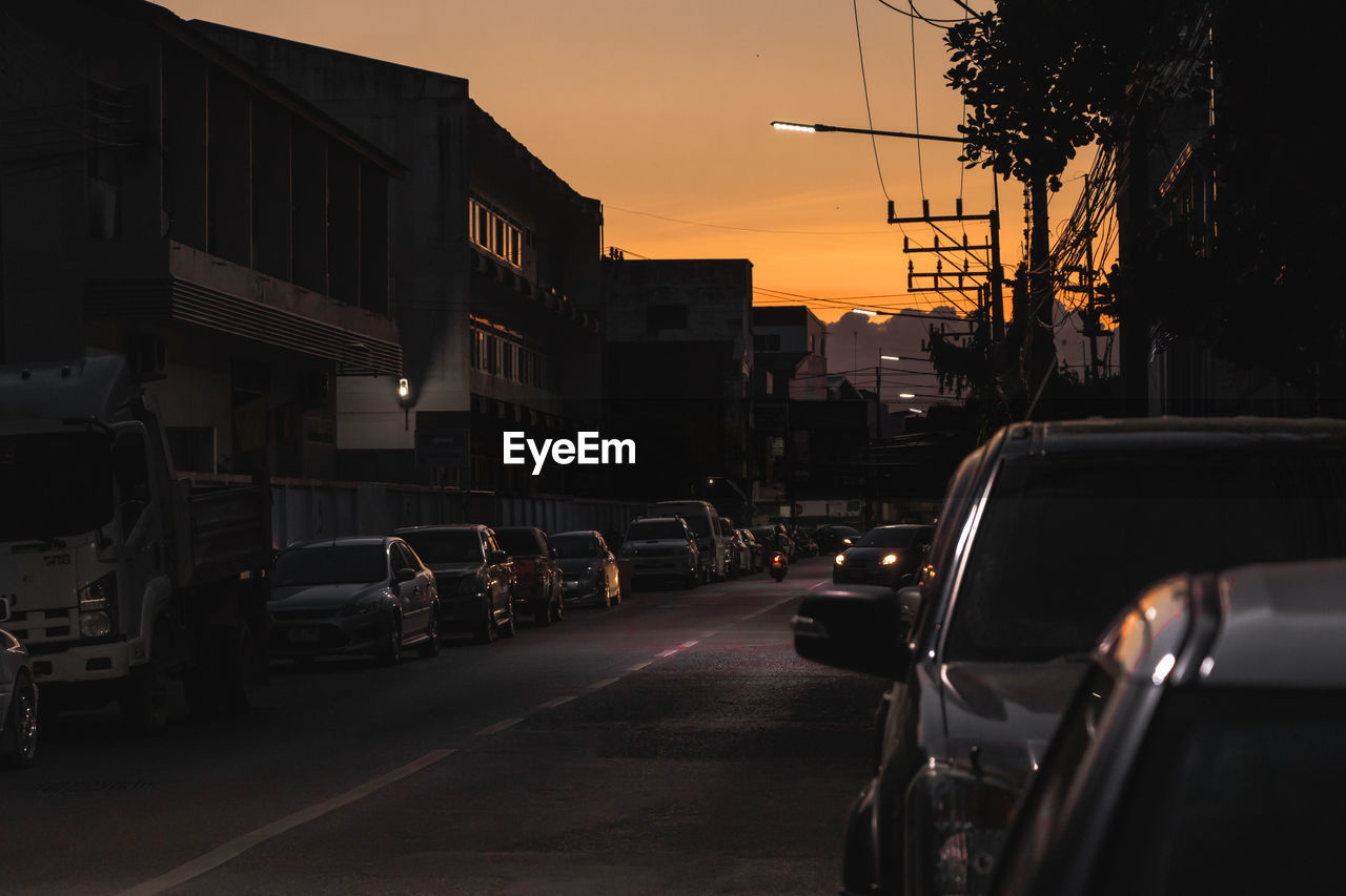 Cars on street by buildings against sky during sunset