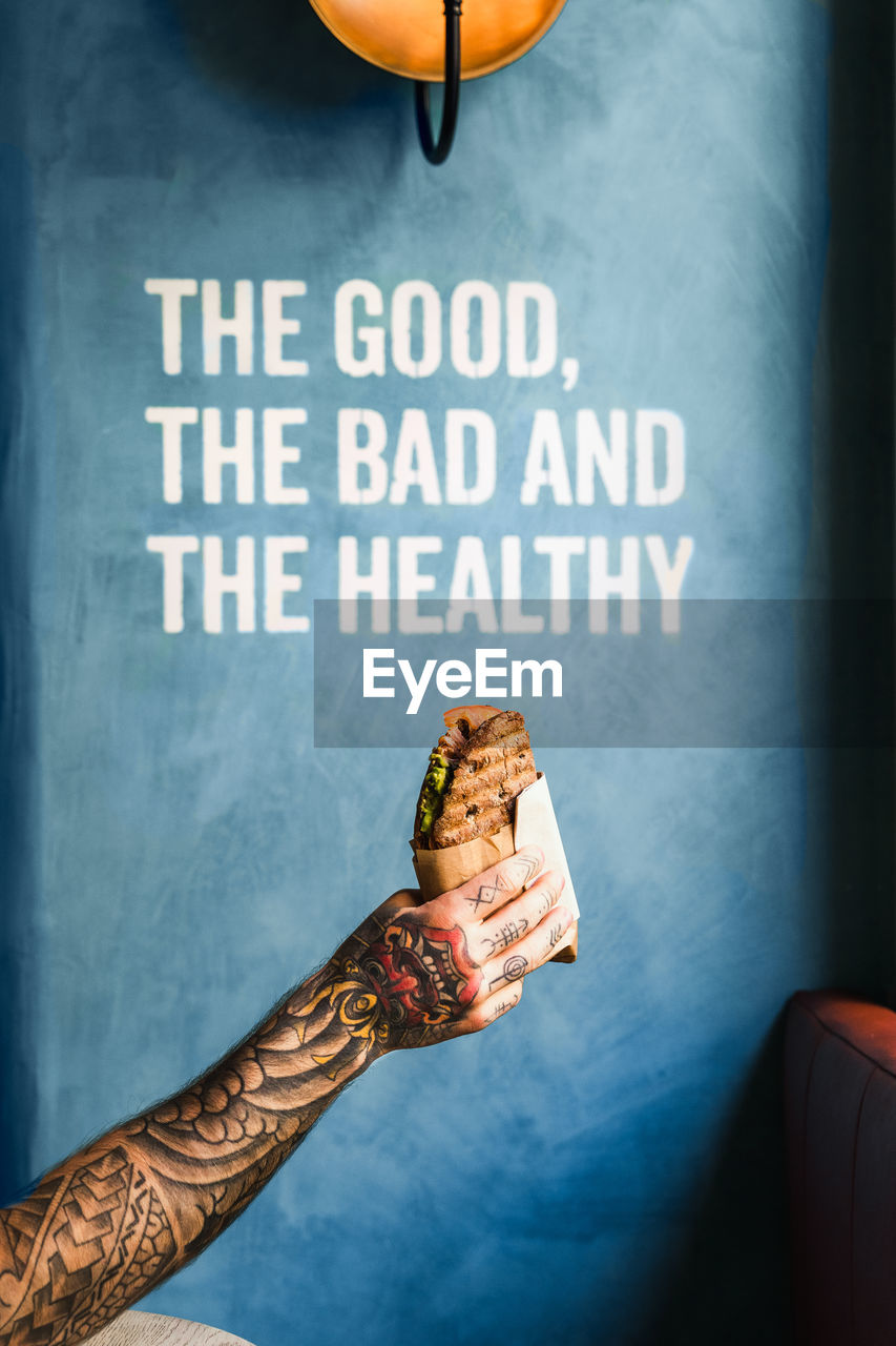 Crop unrecognizable person showing tasty sandwich to go against wall with the good the bad and the healthy inscription person