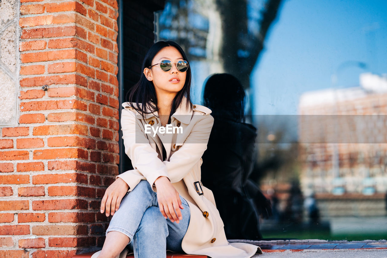 Self assured young asian lady with long dark hair in fashionable outfit in sunglasses and looking away while sitting near brick wall on city street