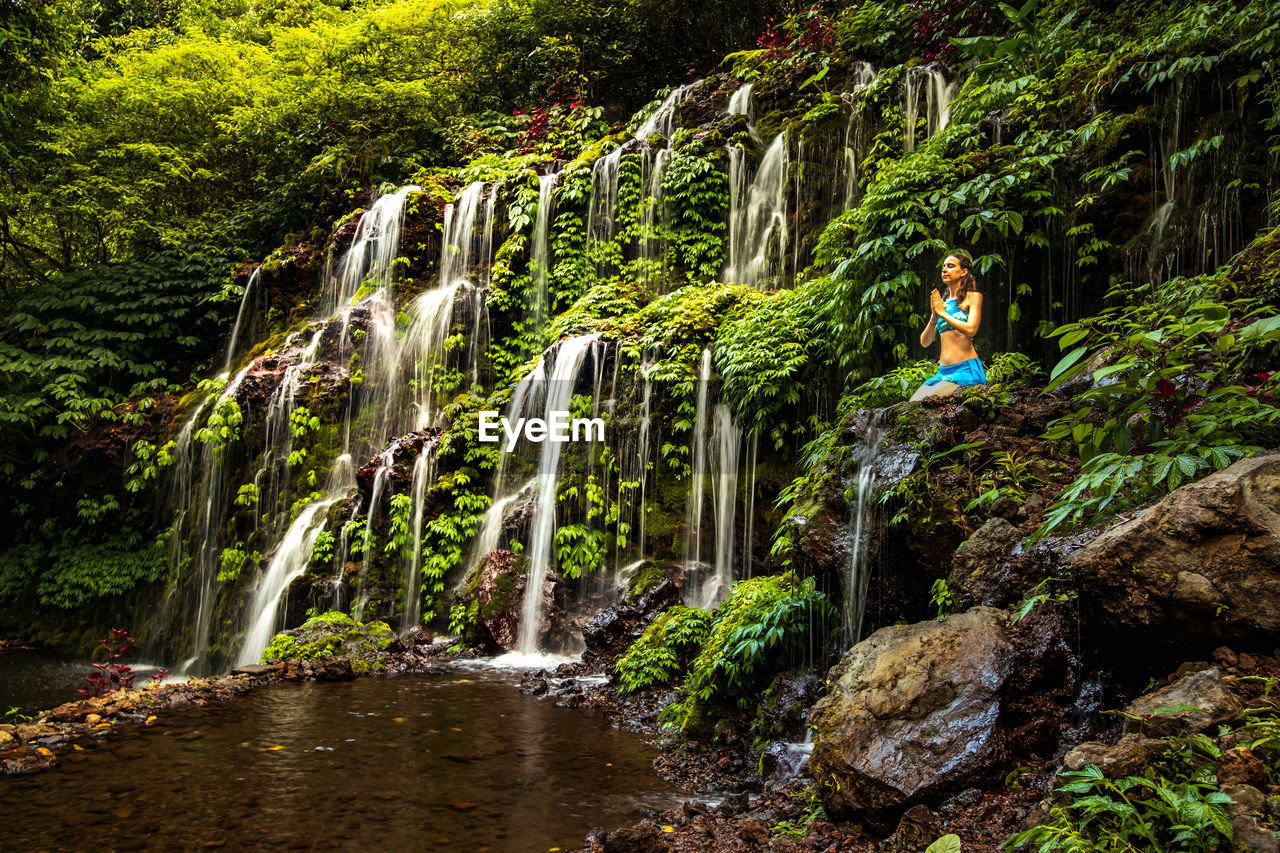 Full length of woman sitting by waterfall in forest