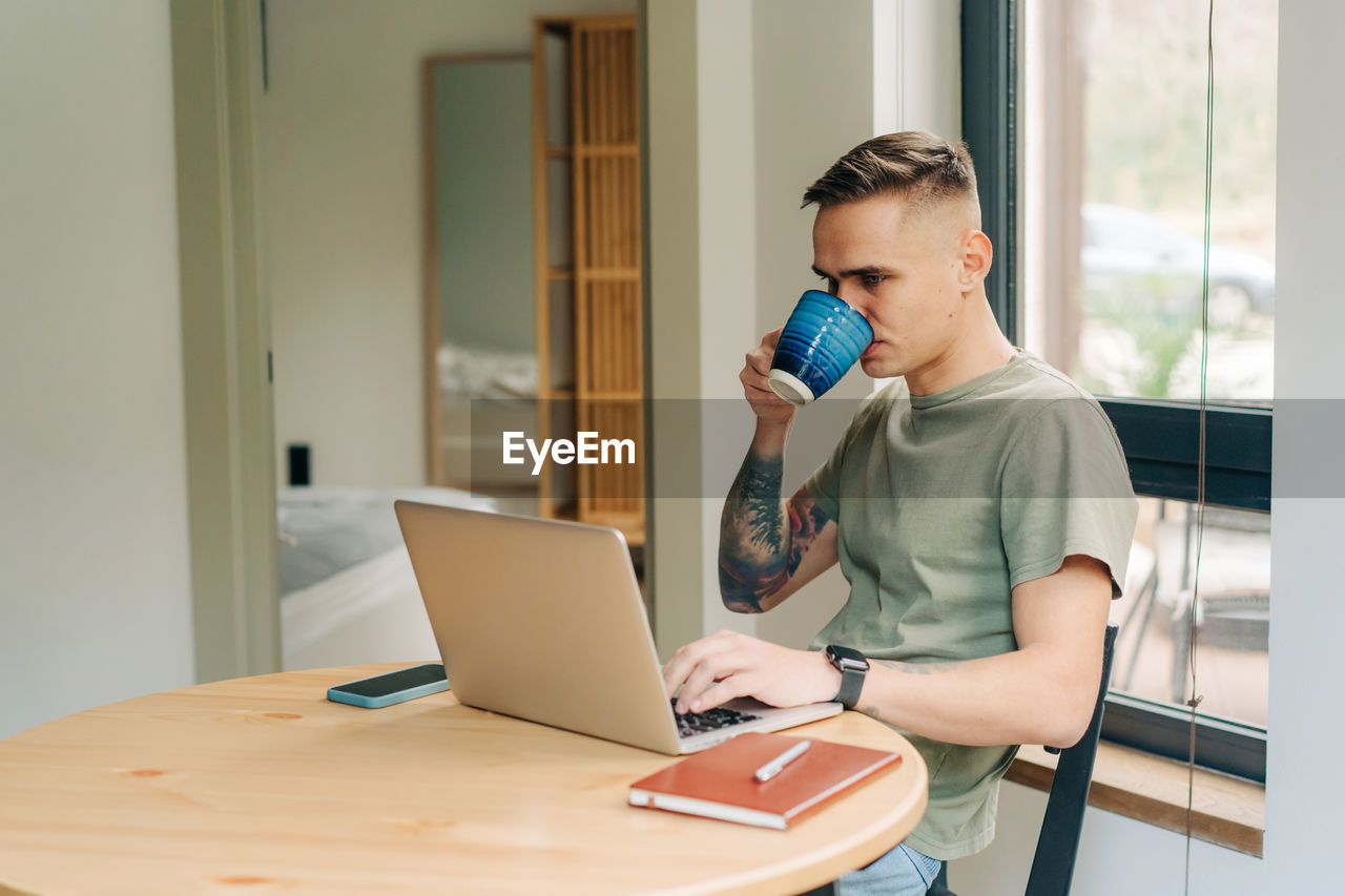 A young man drinks coffee and works remotely on a laptop while sitting at a table in the living room