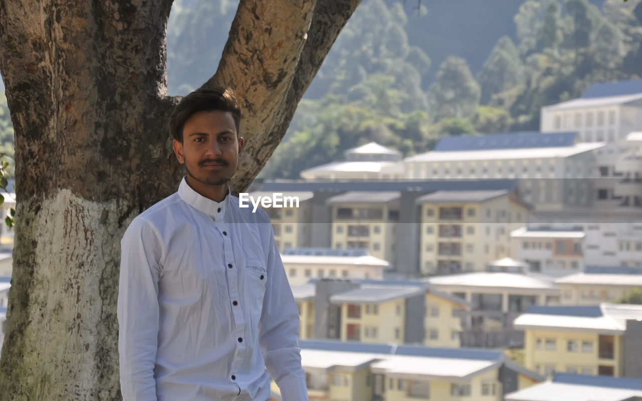 A college student looking at camera standing against tree with college campus in background 