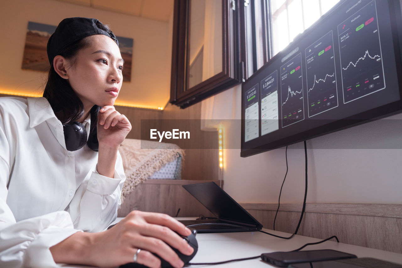 Side view of concentrated asian female working on computer with charts showing dynamic of changes in value of cryptocurrency at convenient workplace