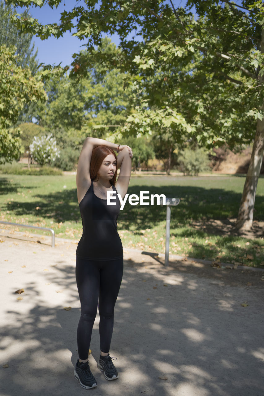 Fitness young redhead woman doing stretching exercise outdoors in public park.