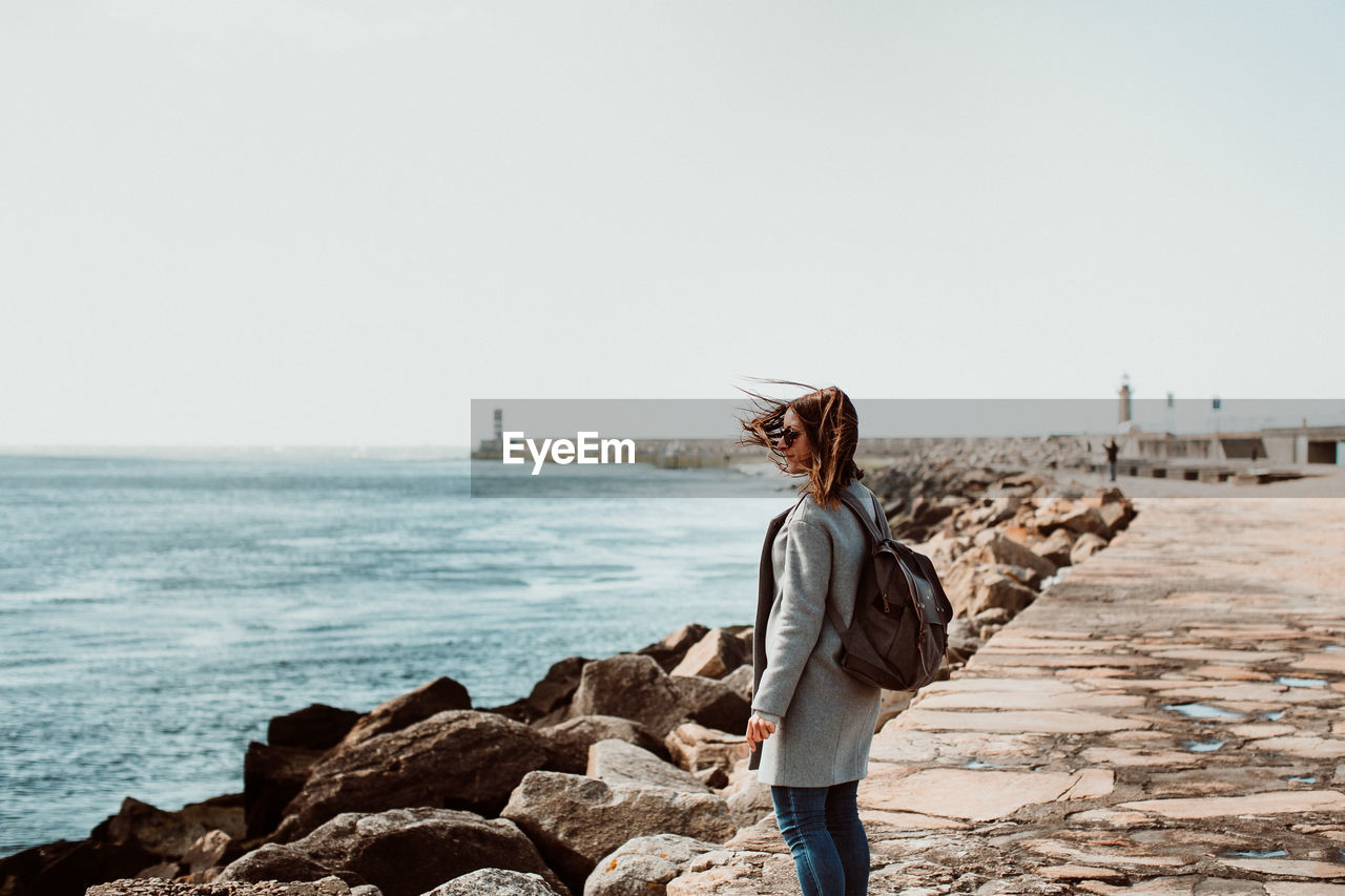 Side view of woman wearing backpack while standing on pier over sea against clear sky