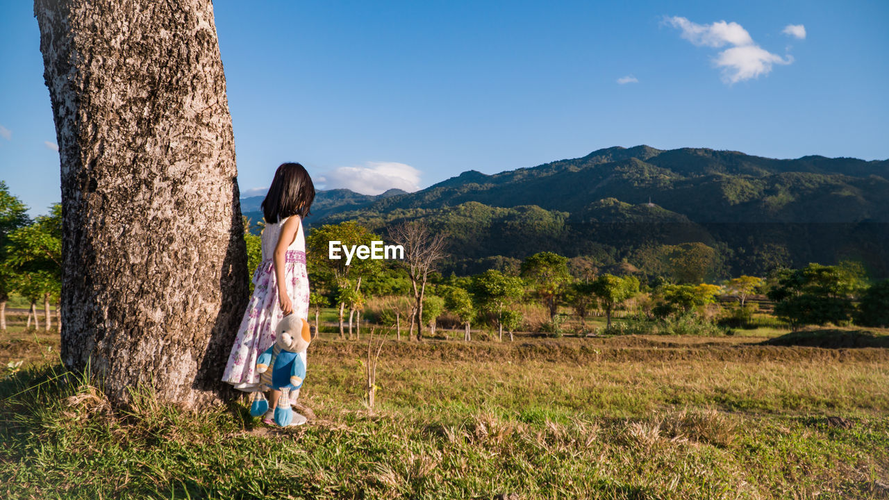 Girl holding toy while standing on grass by tree trunk against mountain and sky