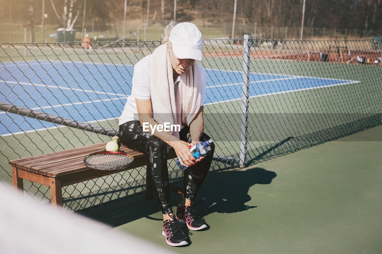 Full length of tired woman sitting on bench against net at tennis court