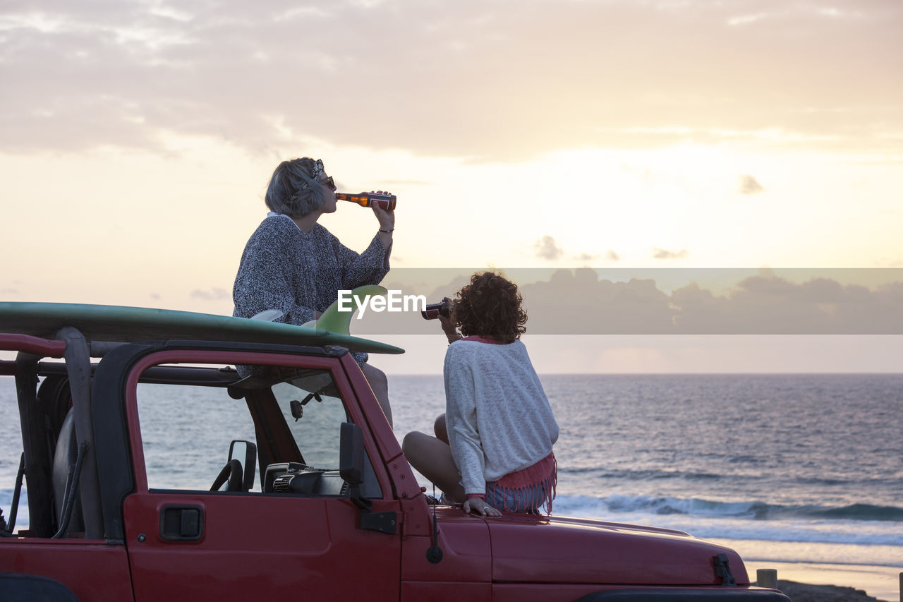 Two surfer girls with sunglasses on their 4x4 car drinking beer