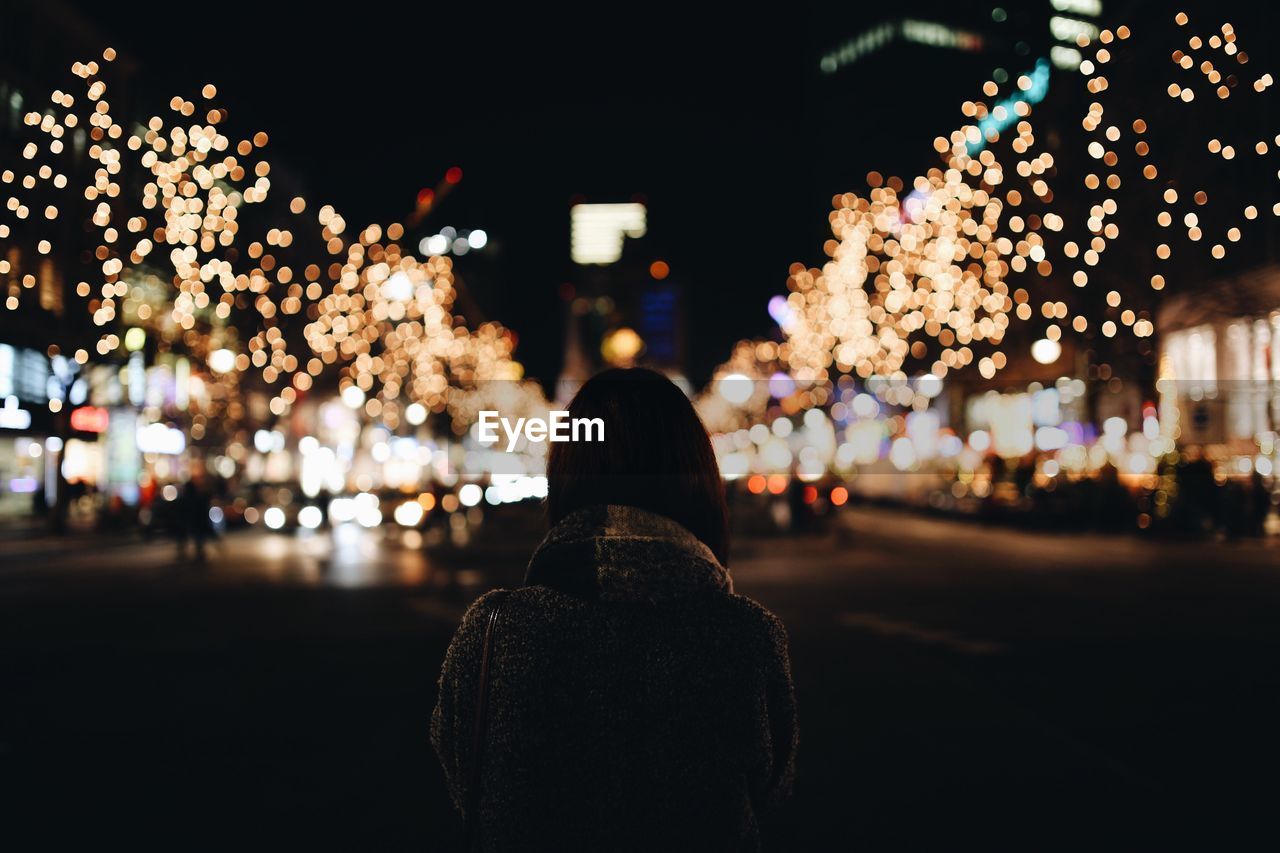 Rear view of woman standing on city street against christmas lights at night