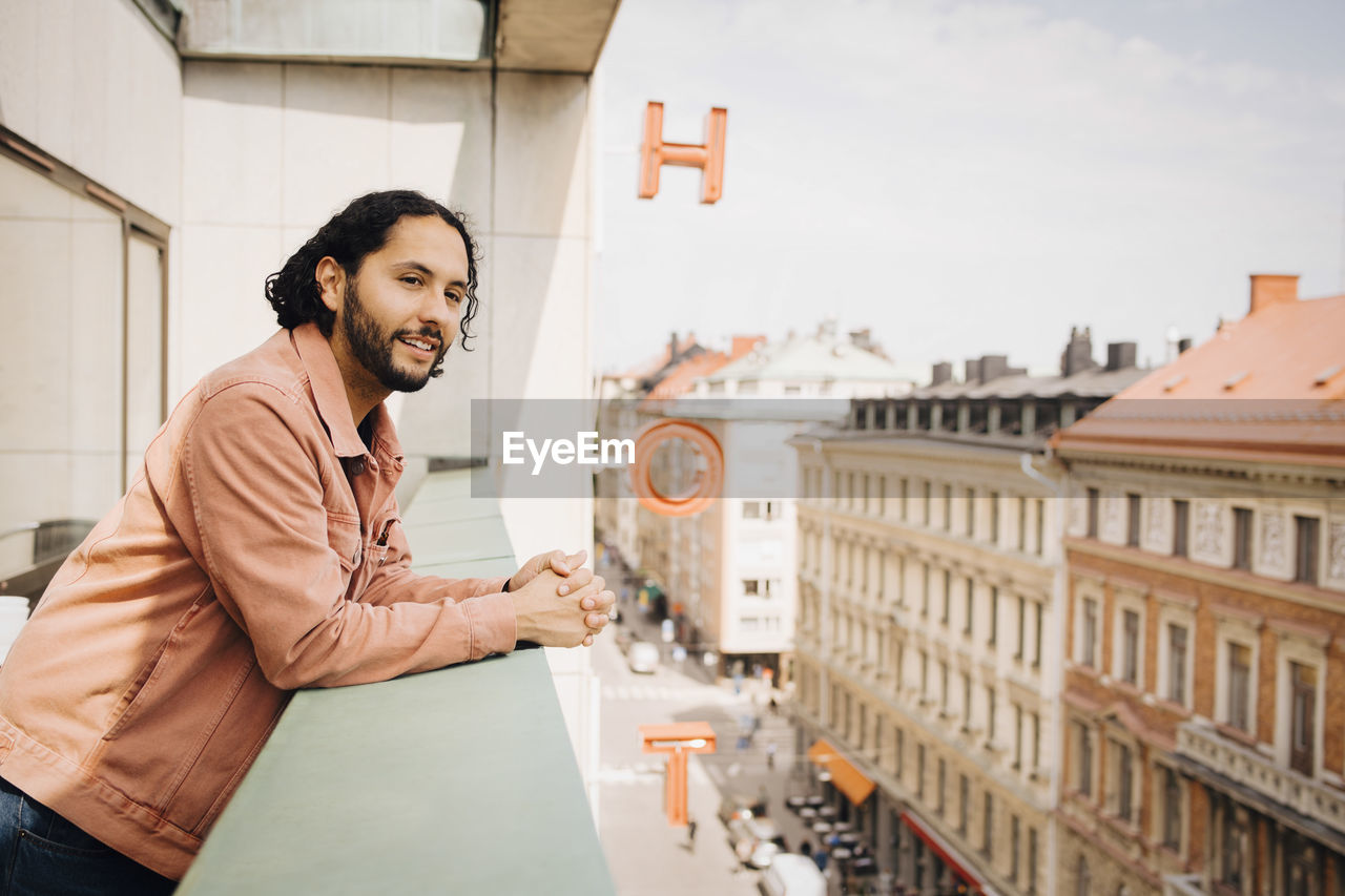 Smiling man looking away while leaning on hotel balcony during sunny day