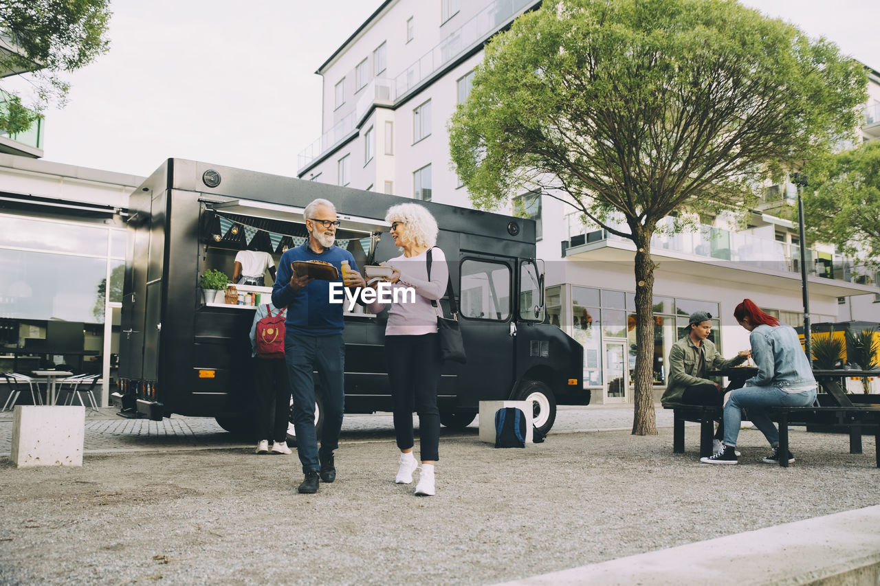 Senior male and female customers with street food walking against commercial land vehicle in city