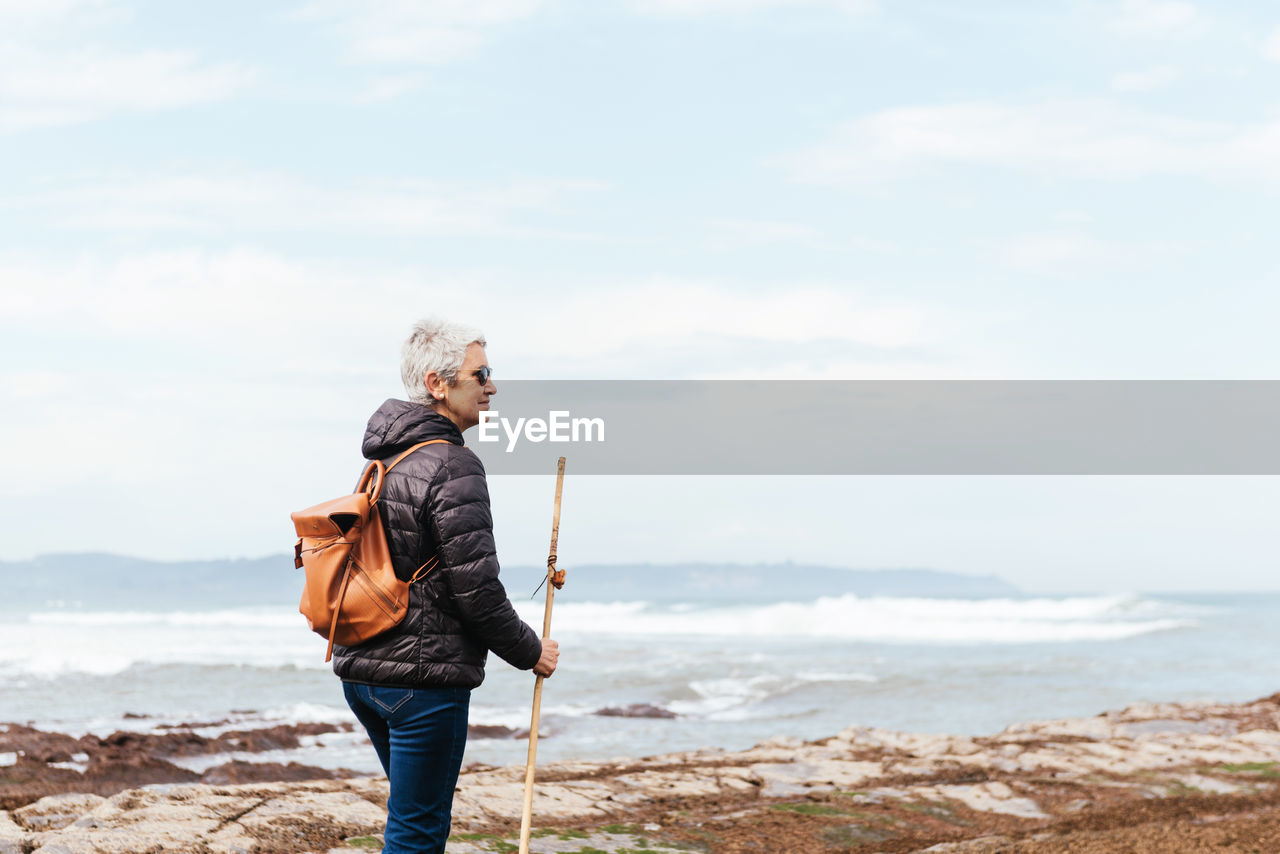 Side view of elderly female backpacker with trekking pole strolling on boulders against stormy ocean under cloudy sky