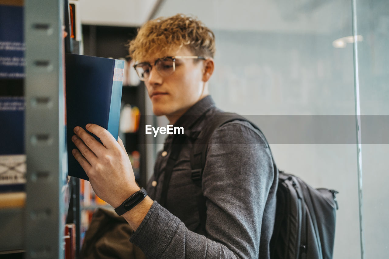 Side view of young man wearing eyeglasses searching book in library at university