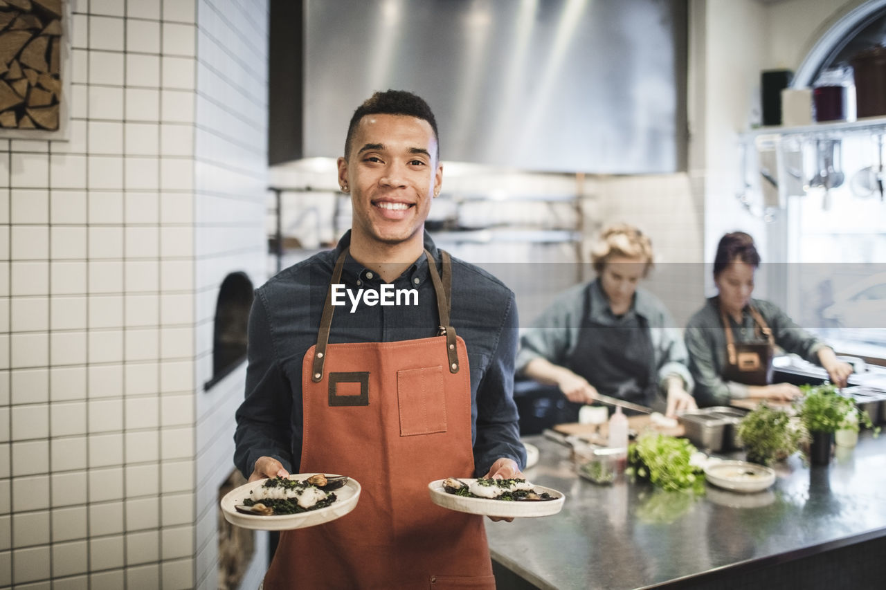 Portrait of smiling waiter holding fresh food plates against female chefs in restaurant kitchen