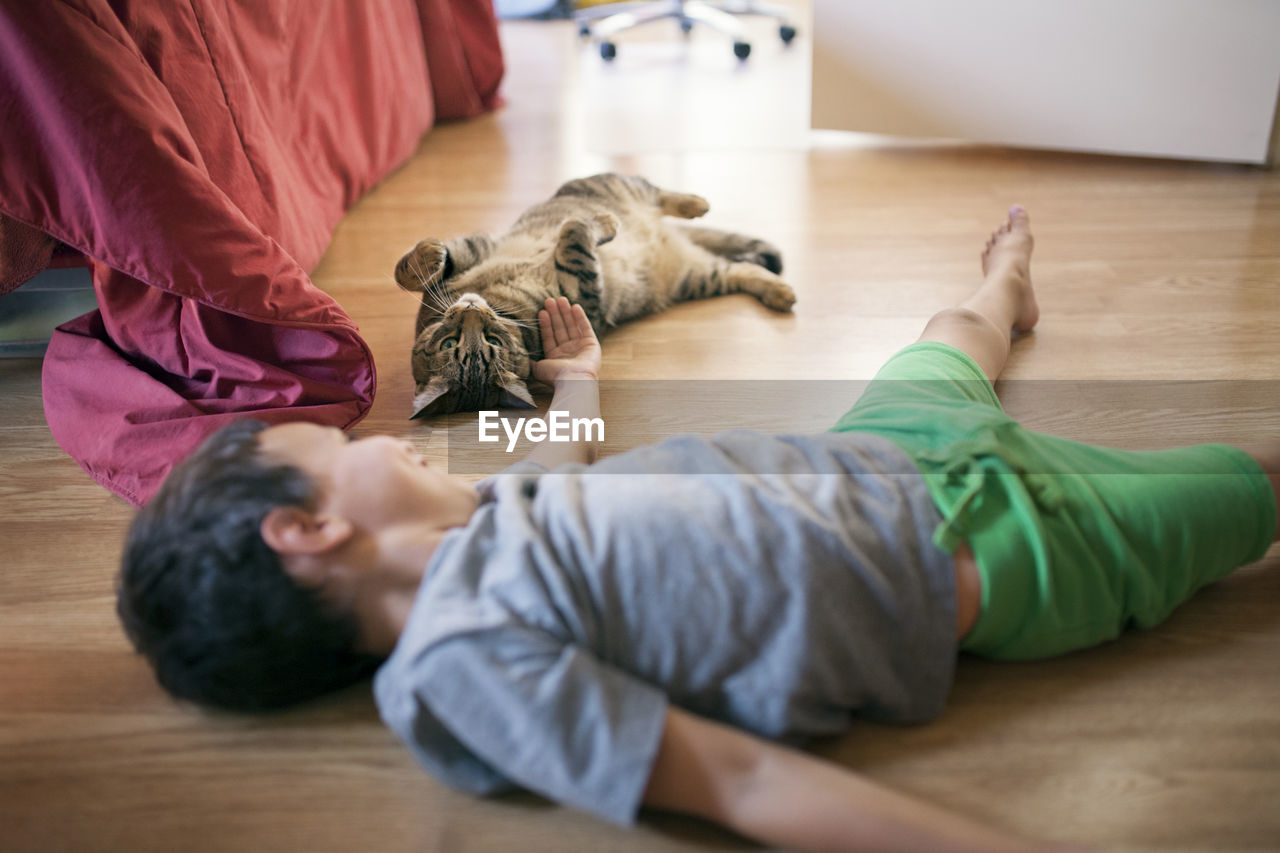 Little boy playing with cat while lying on hardwood floor at home