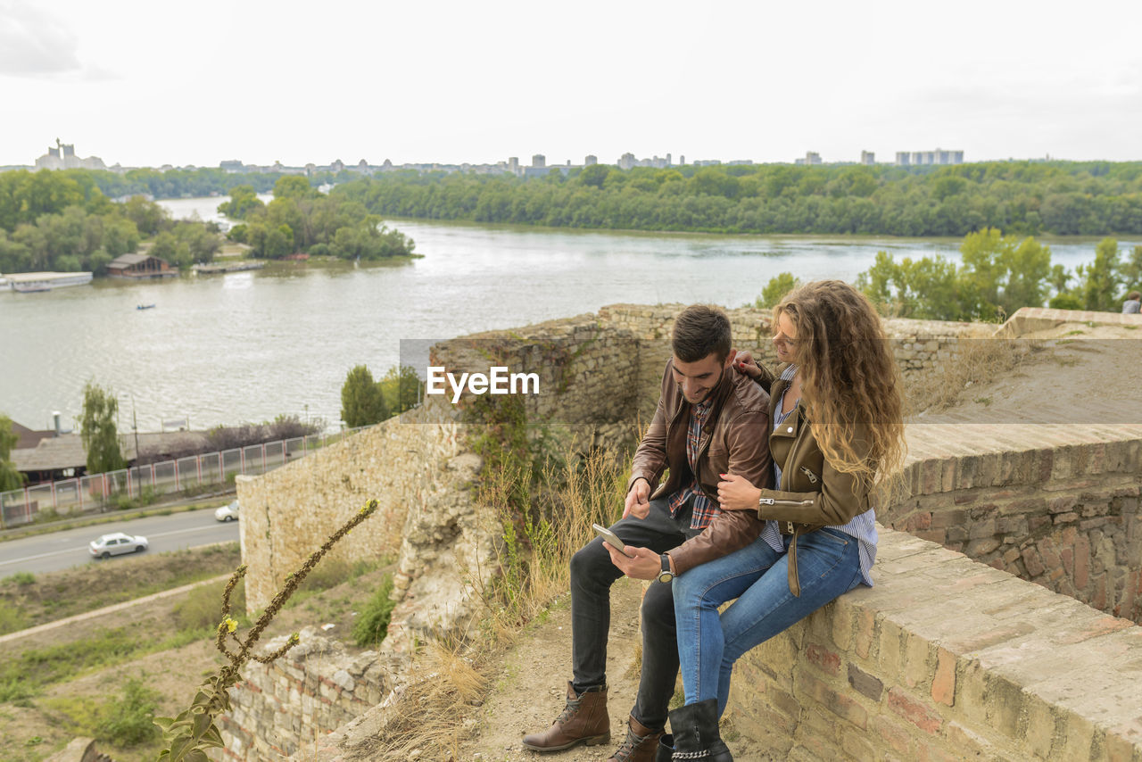 Young couple using mobile phone while sitting on retaining wall