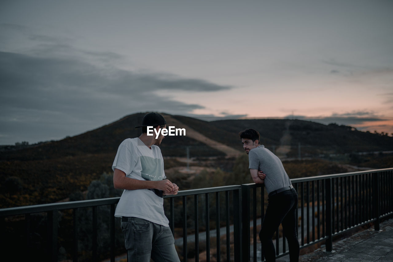 PEOPLE STANDING ON RAILING AGAINST MOUNTAIN