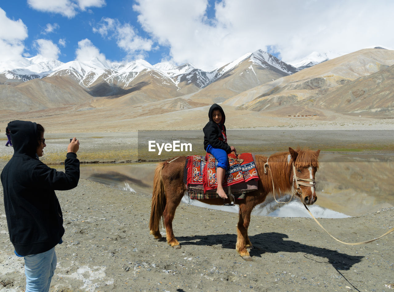 Teenage girl photographing sister sitting on horse against mountains