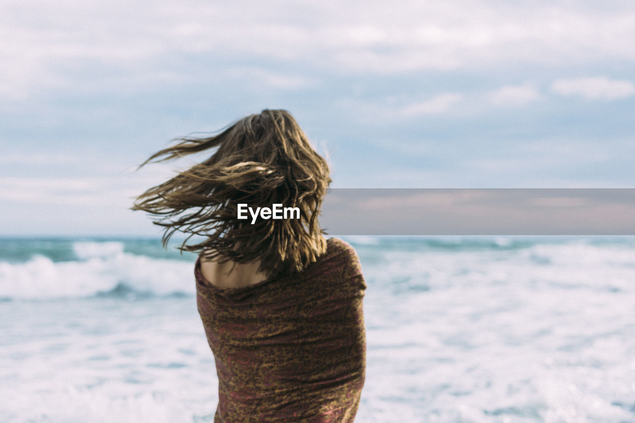 Rear view of mid adult woman with scarf standing at beach against cloudy sky