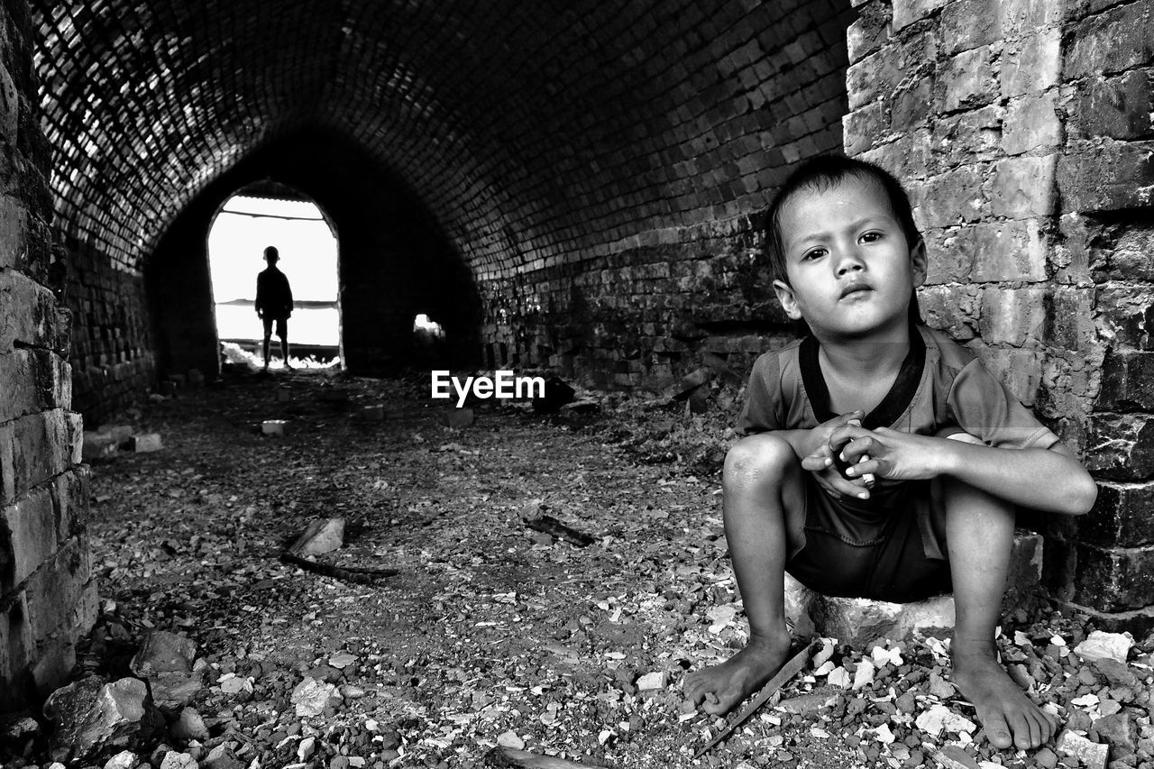 Portrait of boy sitting against brick wall with brother standing in background