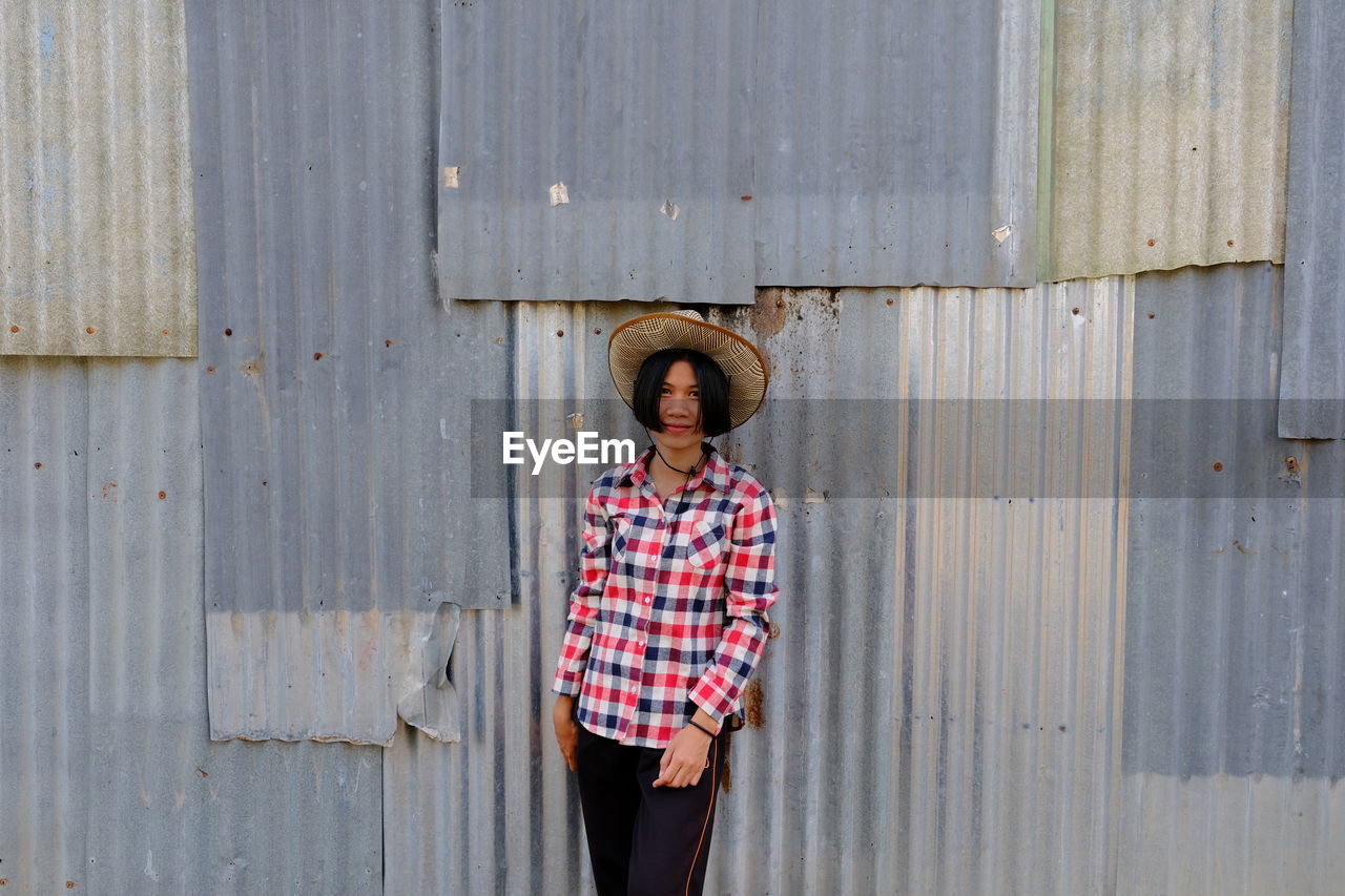 Portrait of woman in hat standing against corrugated iron