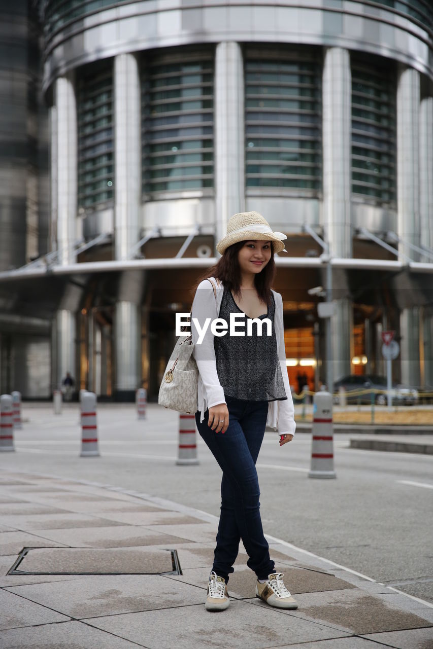 Portrait of young woman standing on sidewalk against modern building in city