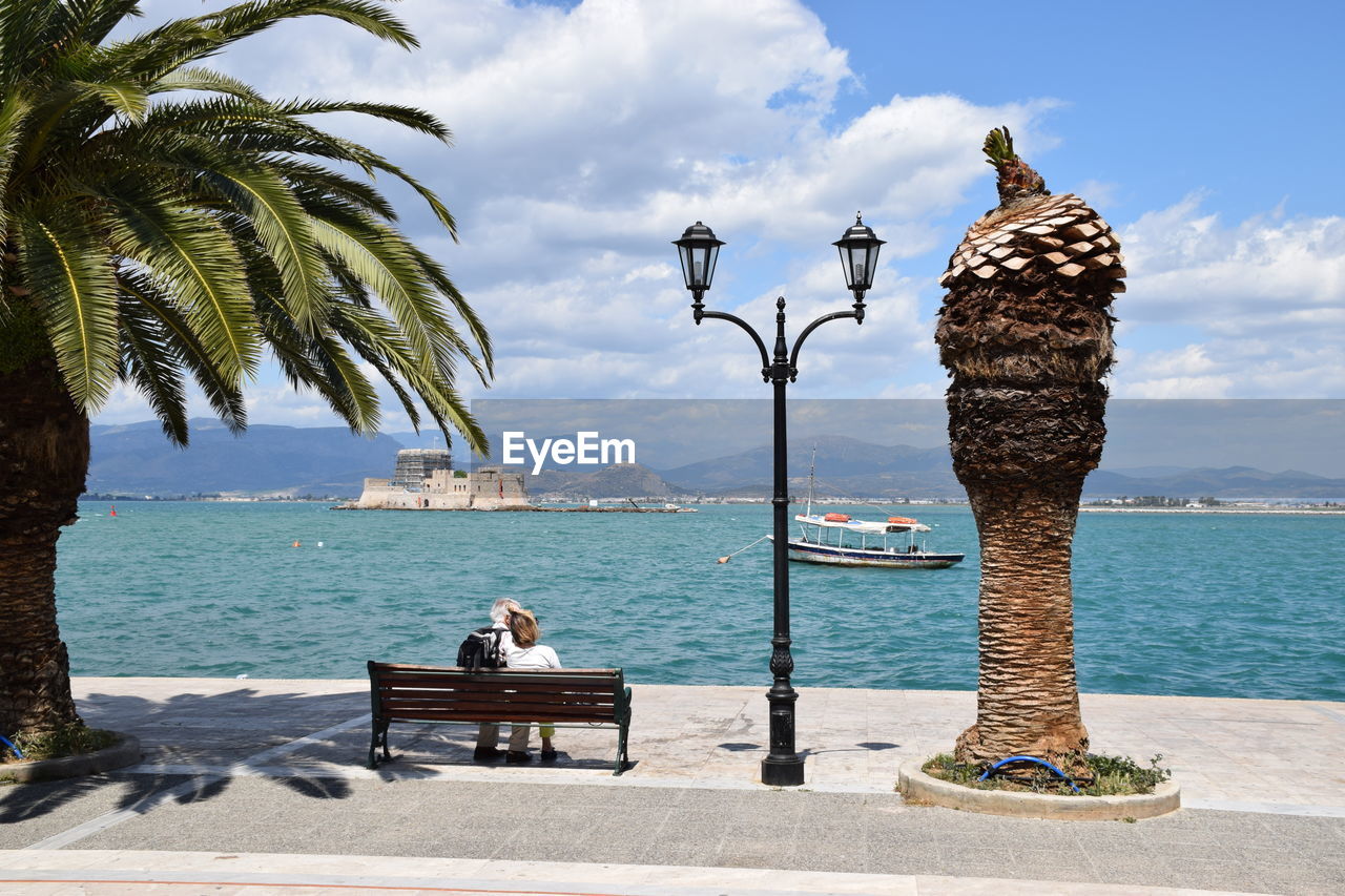 Rear view of people sitting on bench at sea shore by palm trees