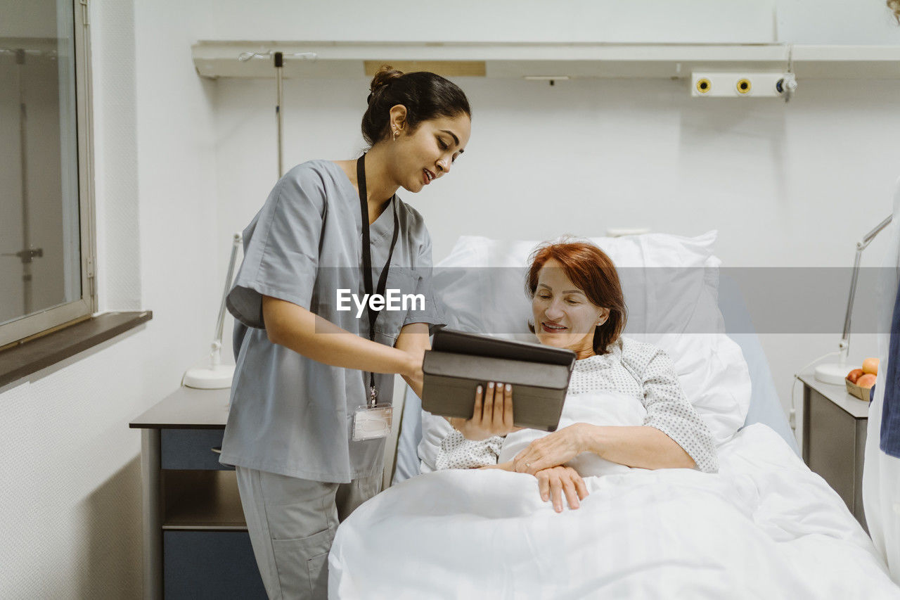 Smiling nurse showing digital tablet to senior female patient lying on bed in hospital