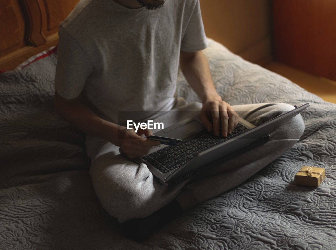 Young caucasian man in gray sweatpants and a t-shirt doing online shopping sitting on the bed