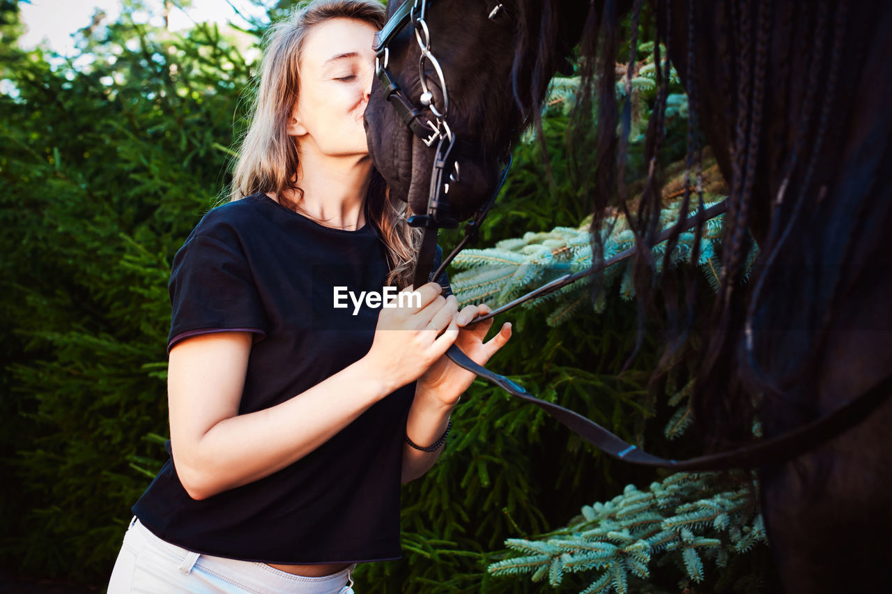 Young woman kissing horse while standing by trees