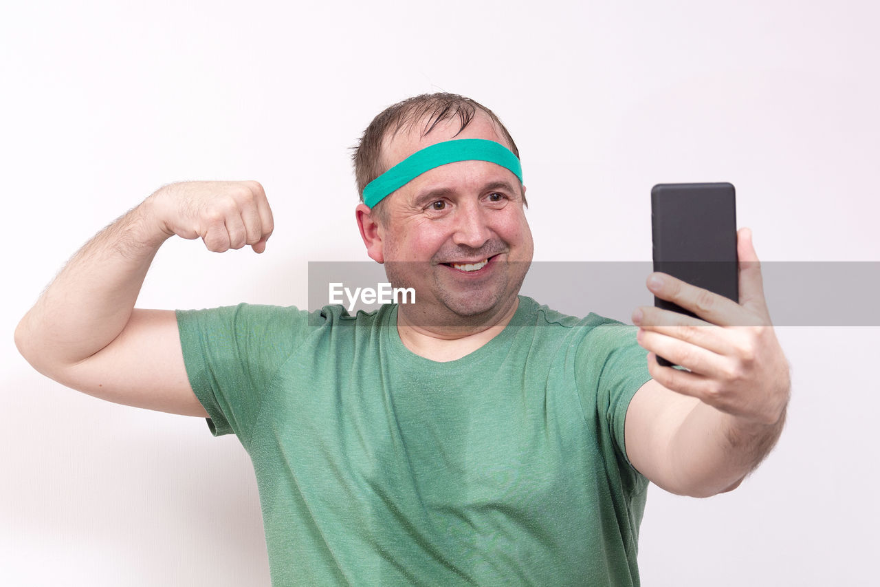 A funny fat man in a green bandana and t-shirt takes a selfie of his right bicep.