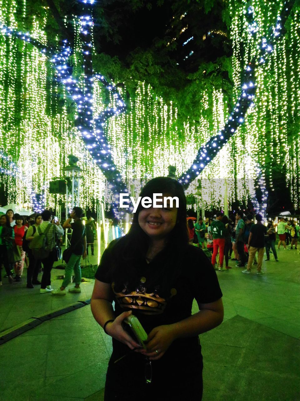 PORTRAIT OF SMILING YOUNG WOMAN STANDING BY ILLUMINATED TREE