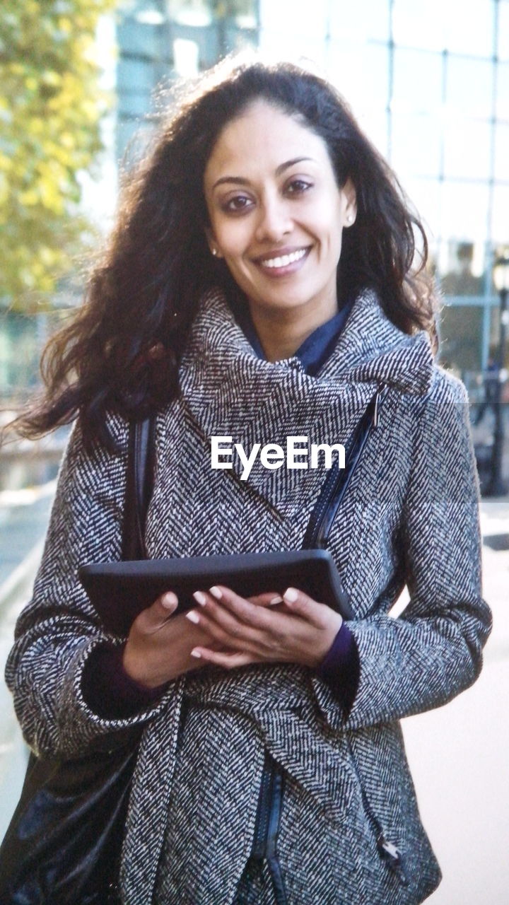 Portrait of smiling woman with digital tablet standing against building