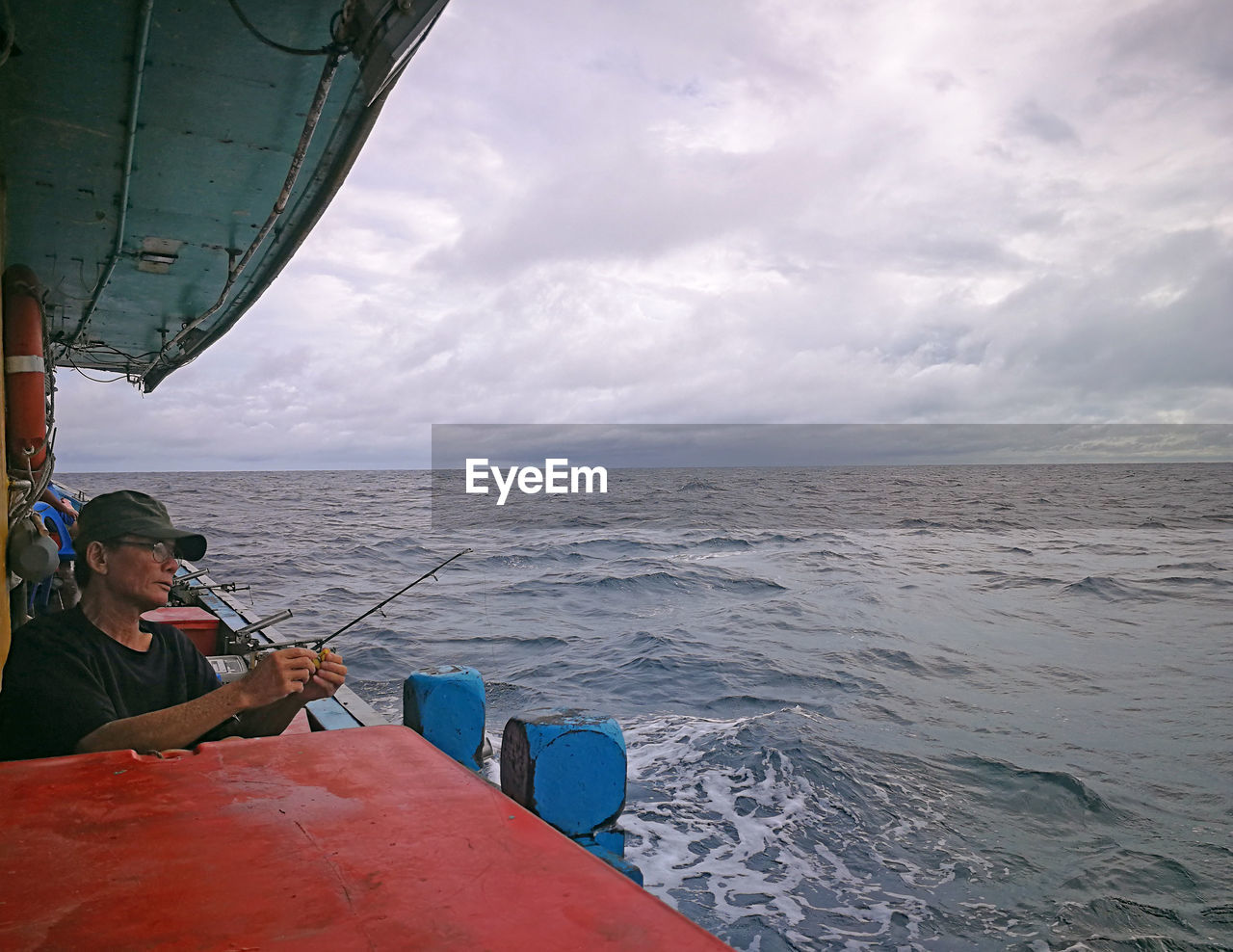 Senior man fishing on boat in sea against cloudy sky
